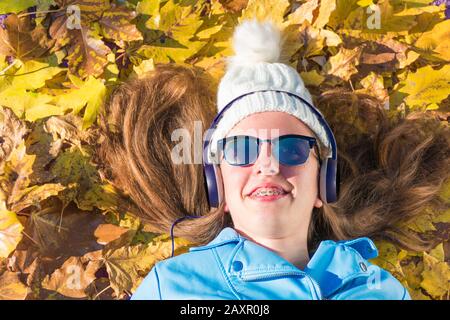 la ragazza adolescente che indossa occhiali da sole e cappuccio è sdraiata sul pavimento coperto da foglie gialle mentre si gode ascoltare musica con le cuffie. Il brun Foto Stock