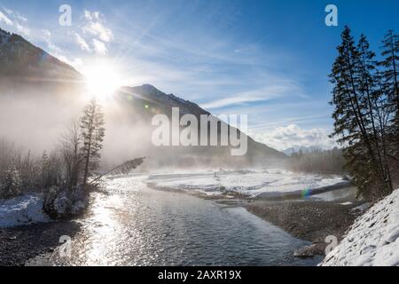 Sole e Sunstar sull'Isar All'Autunno nel Karwendel nel mezzo dell'inverno, il fiume innevato riflette la luce del sole e alcuni alberi di abete si trovano sul Foto Stock