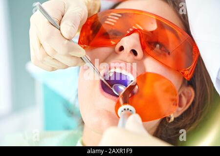 Ragazza bambino al medico. Il dentista posiziona un riempimento su un dente con la lampada di polimerizzazione dentale in cavità orale. Su sfondo clinico Foto Stock