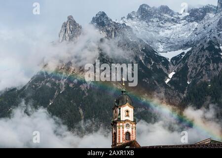 Arcobaleno sopra la torre della chiesa di Mittenwald nel Karwendel bavarese. Sullo sfondo le montagne del Karwendel, parzialmente innevate. Foto Stock