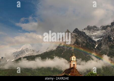 Arcobaleno sopra la torre della chiesa di Mittenwald nel Karwendel bavarese. Sullo sfondo le montagne del Karwendel, parzialmente innevate. Foto Stock