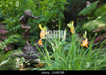 Statuette in metallo di bronzo di giovane ragazzo in piedi su una lumaca e sfumato arancione Hemerocallis - fiori di giorno nel giardino di campagna cortile in estate. Foto Stock