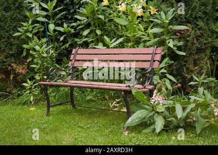 Legno marrone e arrugginito nero ghisa seduta panca in un giardino paesaggistico residenziale cortile in estate. Foto Stock