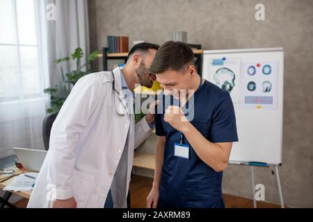 Bearded medico che dice qualcosa di divertente onn l'orecchio al suo collega Foto Stock