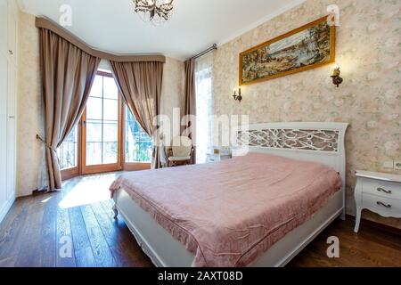 Camera classica in toni beige. Una grande finestra dove il sole splende. Tende scure spesse. Un grande letto bianco con copriletto rosa e un motivo curvo Foto Stock