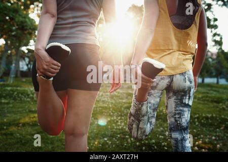 Primo piano di due giovani donne sportive che allungano le gambe nella giornata di sole nel parco - due donne multirazziali che allungano i loro quad prima di una corsa Foto Stock