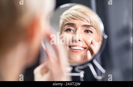 Donna Di Mezza Età Che Guarda Nello Specchio Che Tocca Le Guance Lisce In Bagno Foto Stock