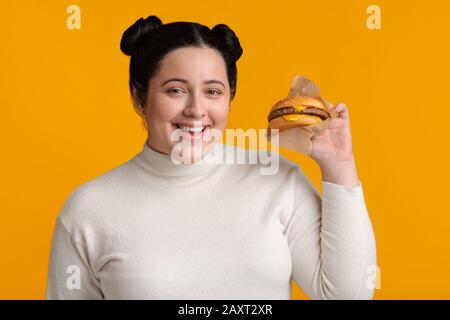 Ragazza Di Sovrappeso Che Posa Con Hamburger Di Manzo Nelle Mani Su Sfondo Giallo Foto Stock