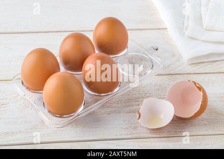 Cinque uova di pollo crude in un vassoio di plastica trasparente e guscio d'uovo vuoto vicino ad esso su un tavolo di legno bianco. Cucina per pasqua con uova marroni. Vista dall'alto. Foto Stock