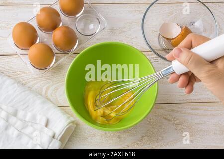 Donna mano che frusta un uovo in una ciotola verde. Cinque uova di pollo marroni in un vassoio di plastica trasparente su un tavolo di legno bianco. Cucina per pasqua. Foto Stock
