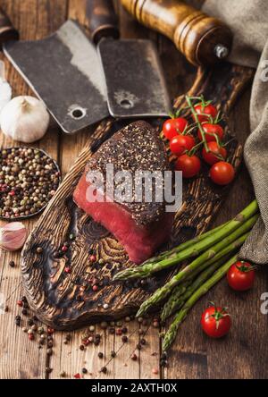 Fetta di manzo crudo fesa giunto con il sale e il pepe sul tagliere di legno con carne hatchet, aglio e punte di asparagi su legno tavolo da cucina. Foto Stock