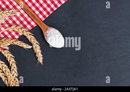 Cucchiaio di legno con farina, spighe di grano e panno rosso a scacchi sul tavolo nero Foto Stock