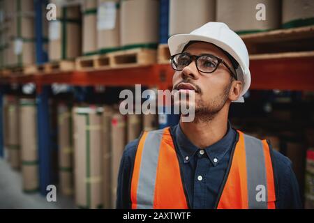 Supervisore maschile di magazzino in uniforme e il pensiero casco in piedi in fabbrica guardando lontano Foto Stock