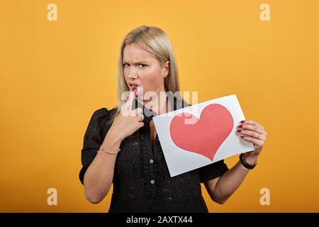 la donna arrabbiata tiene un pezzo di carta con il cuore rosso in mano infila un dito in bocca Foto Stock