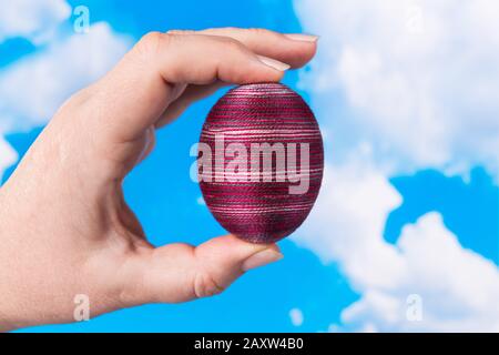 Originale uovo di Pasqua striato in mano femminile su sfondo cielo blu primavera. Guscio di pollo rosso decorato da filato di cotone sottile incollato in dita umane. Foto Stock