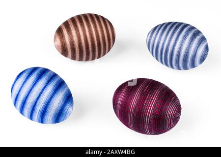 Quattro uova di Pasqua striate decorative su sfondo bianco. Primo piano di gusci d'uovo di colore blu, rosso o marrone decorati con filo per cucire di cotone incollato. Foto Stock