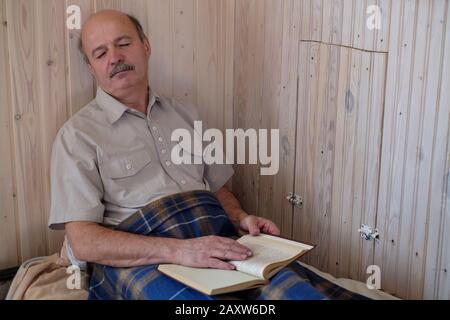 Uomo anziano che dorme sul divano interno riposante durante il giorno e libro di lettura Foto Stock