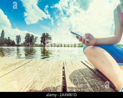 Una ragazza con un telefono in mano siede sulla superficie di legno vicino alla superficie dell'acqua. Scatto dal basso. Fotocamera grandangolare professionale. Foto Stock