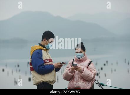 I residenti cinesi locali indossano maschere facciali per prevenire il nuovo coronavirus e la polmonite usano i loro smartphone presso il West Lake di Hangzhou City Foto Stock