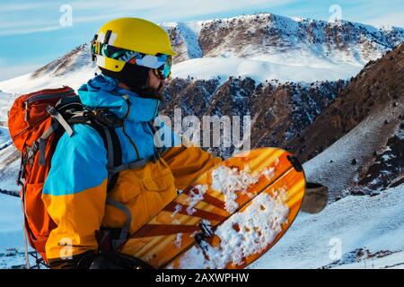 Un uomo con uno snowboard nelle sue mani in montagna. Sta considerando un pendio. Montagne del Kirghizistan Foto Stock