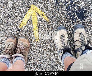 Gli stivali di due camminatori pellegrini si trovano accanto a un Camino a Santiago de Compostela in Spagna. Freccia gialla dipinta sulla strada lastricata. Foto Stock