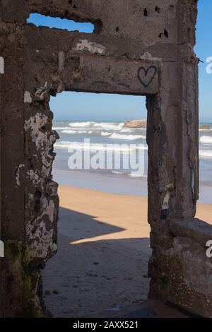 Particolare di un edificio abbandonato, con un cuore spruzzato sulla parete rotta. Spiaggia e oceano Atlantico sullo sfondo. Essaouira, Marocco. Foto Stock