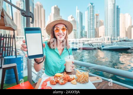 Asian girl mangia la cucina di pesce che guarda ai grattacieli e al porto di Marina a Dubai e mostra uno schermo vuoto del suo smartphone. Concetto di app per ca Foto Stock