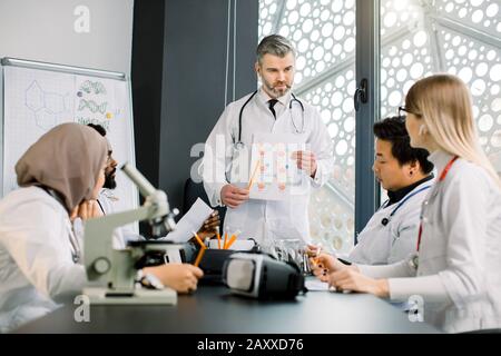 Bel medico di mezza età che spiega le informazioni per i suoi studenti alla classe. Il medico supervisore maschio sorridente mentre parla con i suoi giovani Foto Stock