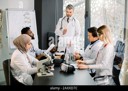 Multietnico quattro giovani medici e il loro professore di insegnante che hanno una discussione durante la conferenza con documenti al tavolo nel moderno ufficio Foto Stock