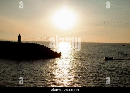 Una piccola barca che arriva al riparo di un interruttore d'onda. Il riflesso del sole che tramonta splende sulla superficie del mare. Foto Stock