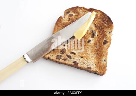 Spalmare il burro su una fetta di pane tostato fresco e caldo con uvetta usando un coltello a manico d'osso, vista ad alto angolo su bianco Foto Stock