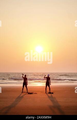 due uomini irriconoscibili che si esercitano al tramonto tenendo due pesi piccoli su un'ampia spiaggia di sabbia vuota aperta, sono in piedi con le loro braccia in estetc Foto Stock