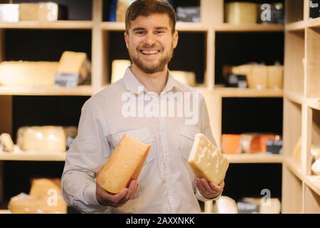 Un bell'uomo con bearded tiene in mano diversi tipi di formaggio invecchiato nel negozio di formaggi Foto Stock