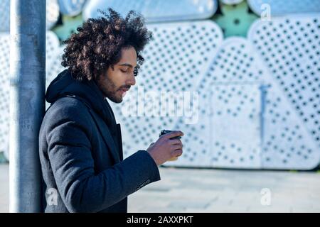 Vista laterale di un Hipster afro uomo in piedi con caffè da asporto, sorridente piacevolmente, in strada della città. Generazione millenaria Foto Stock
