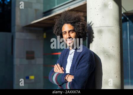 Un giovane e bello afroamericano alla moda si trova vicino a un pilastro. Uno studente in jeans e una giacca blu con un'acconciatura alla moda. Moda giovane c Foto Stock