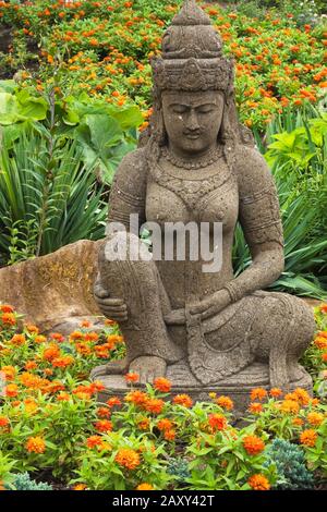 Pietra marrone scultura dea indonesiana in arancio Zinnia fiore letto nel giardino Zen in estate al giardino Route des Gerbes d'Angelica. Foto Stock