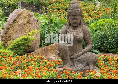 Pietra marrone scultura dea indonesiana in arancio Zinnia fiore letto nel giardino Zen in estate al giardino Route des Gerbes d'Angelica. Foto Stock