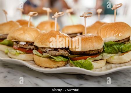 Hamburger con tortino di manzo, pomodoro fresco, lattuga, cipolla fritta e formaggio fuso tenuto con un spiedino di legno, servito su un piatto bianco di ceramica. Foto Stock