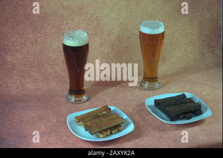 Due bicchieri alti con birra chiara e scura su sfondo marrone con circa due piatti con crostini fritti. Primo piano. Foto Stock