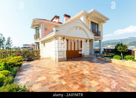 La facciata di un cottage classico bianco a due piani con giardino frontale. Porta d'ingresso scolpita, cancello auto, piastrelle beige, alberi. Foto Stock