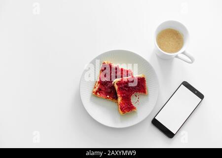 La colazione è a tavola. Una tazza di caffè e toast con marmellata di ciliegie accanto al telefono. Foto Stock