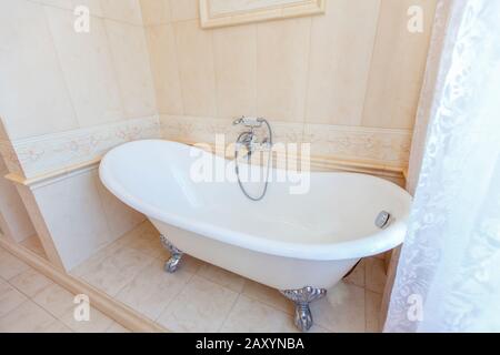 Bagno classico in toni beige. Vasca da bagno curva. Grande piastrella beige. Piastrelle bianche sul pavimento. Lampadario ritorto, elegante e costoso impianto idraulico Foto Stock