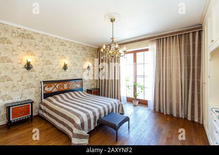 Elegante camera da letto costosa con un grande letto bianco con un copriletto a righe. Un'alta testata con un motivo rosso, lampade a parete intrecciate e un lampadario. B Foto Stock