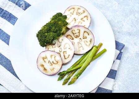 Verdure al vapore in una piastra bianca su un tavolo blu. Melanzana, Broccoli asparagi. Foto Stock