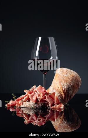 Il Prosciutto con ciabatta, vino rosso e il timo su un nero sfondo riflettente. Foto Stock