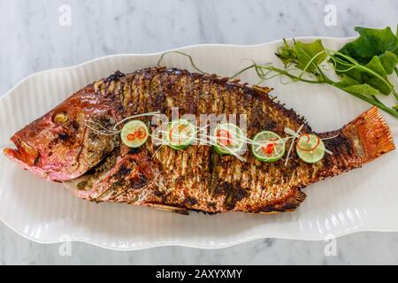 Pesce intero alla griglia servito su un piatto bianco di ceramica. Decorato con lime e peperoncino tagliato. Vista dall'alto. Foto Stock