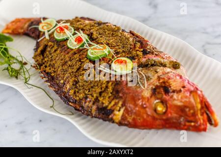 Pesce intero alla griglia servito su un piatto bianco di ceramica. Decorato con lime e peperoncino tagliato. Vista dall'alto. Foto Stock