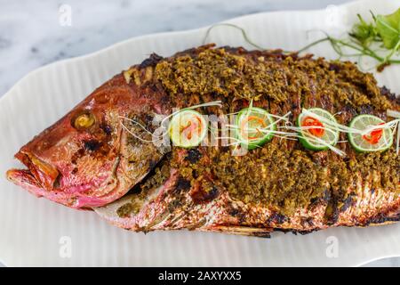 Pesce intero alla griglia servito su un piatto bianco di ceramica. Decorato con lime e peperoncino tagliato. Vista dall'alto. Foto Stock