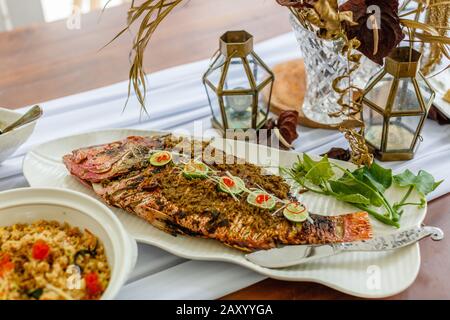 Pesce intero alla griglia servito su un piatto bianco di ceramica. Decorato con lime e peperoncino tagliato. Riso e verdure sul lato. Foto Stock