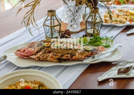 Pesce intero alla griglia servito su un piatto bianco di ceramica. Decorato con lime e peperoncino tagliato. Riso e verdure sul lato. Foto Stock